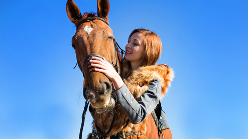 A girl with red hair stands near a brown horse, strokes her nose with her hand, holds a bridle and smiles.